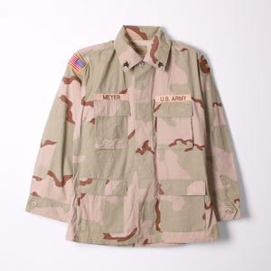Könnte beinhalten: Wüsten-Tarnjacke mit den Aufnähern "MEYER" und "U.S. ARMY". Mit einem amerikanischen Flaggenaufnäher auf der Schulter. Die Jacke hat vier Vordertaschen und geknöpfte Manschetten. Die Farbpalette umfasst Beige, Braun und Grün.