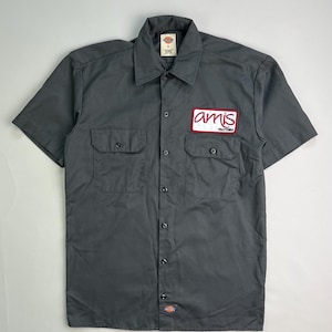 May include: A gray short-sleeved work shirt with two chest pockets and a button-down front. The shirt features a red and white patch that reads "amis trattoria". The Dickies brand label is visible on the collar.