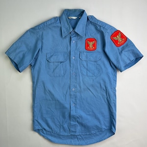 May include: Light blue short-sleeved shirt with two chest pockets and a button-down front. The shirt features two red patches with gold emblems on the sleeves. The shirt is made of a lightweight fabric.
