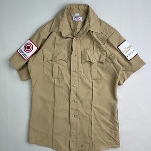 May include: Tan short-sleeve button-up shirt with two chest pockets. Patches on the sleeves read "Marksman" and "Tusca". The shirt has a collared neckline and a button-down front. The shirt is made of a durable, possibly workwear material.