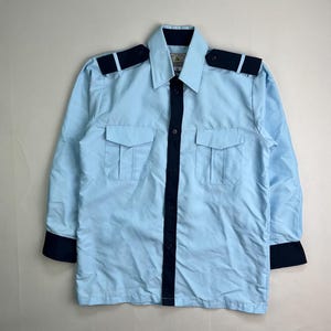 May include: Light blue long-sleeve shirt with two chest pockets, dark blue shoulder epaulets, and dark blue trim on the cuffs and down the front. The shirt has a collared neckline and button closures.