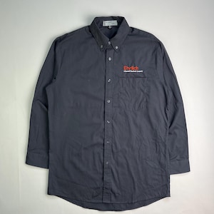 May include: A dark gray button-down shirt with long sleeves. The shirt has a button-down collar and a chest pocket. The word "Ehrlich" is embroidered in red on the chest pocket. The shirt is made of a smooth, woven fabric.