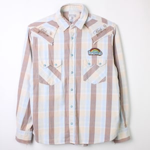 May include: A long-sleeved, button-down shirt with a plaid pattern in brown, blue, and cream. Features two chest pockets and patches with text and a rainbow. The shirt has a Western style with a pointed collar.