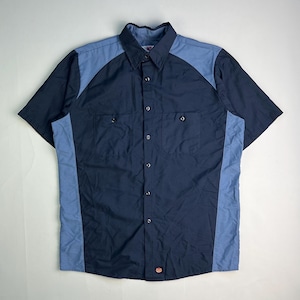 May include: A short-sleeved work shirt in navy blue and light blue. The shirt has two chest pockets, a button-down front, and a collar. The sleeves and side panels are light blue, while the main body is navy blue. A small red logo is visible.