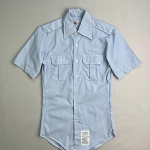 May include: A light blue short-sleeved shirt with two chest pockets and epaulets. The shirt has a button-down front and a collar. There is a small tag at the bottom of the shirt. The shirt is made of a light-weight fabric.
