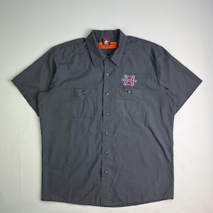 May include: Gray short-sleeve button-up shirt with two chest pockets. Features a red, white, and blue cross graphic on the left chest and a small skull logo on the collar. The shirt has a classic workwear style.