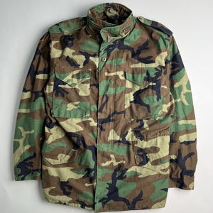 May include: Camouflage pattern jacket in shades of green, brown, and black. The jacket features a button-up front, multiple pockets, and a collared neckline. The military-style jacket is a classic outerwear piece.