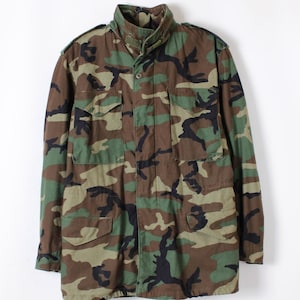 May include: Camouflage pattern jacket in shades of green, brown, and black. The jacket features a high collar, button and zipper closure, and multiple front pockets. A classic military-style outerwear piece.