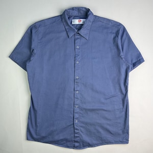 May include: A short-sleeved, button-down shirt in a solid periwinkle blue. The shirt has a classic collar and a small tag near the top button. The shirt is made of a lightweight fabric.
