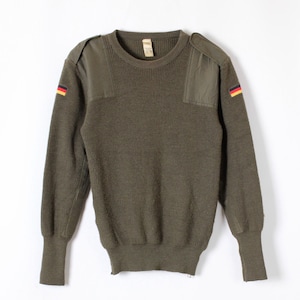 Może przedstawiać: Oliwkowy sweter z naszywkami na ramionach i naszywkami z flagą Niemiec na rękawach. Sweter ma prążkowaną dzianinę i okrągły dekolt. Mankiety i dół są prążkowane. Odzież w stylu vintage militarnym.