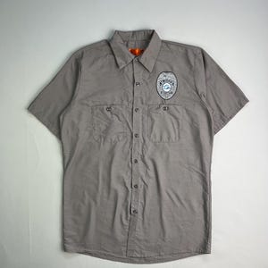 May include: A gray short-sleeved button-down shirt with two chest pockets. A badge-like emblem is on the left chest. The shirt has a classic collar and an orange tag at the neck.