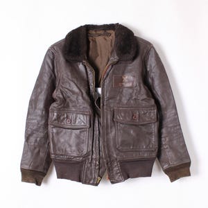 May include: A brown leather bomber jacket with a fur collar. The jacket has two front pockets with button closures, a zipper closure, and a patch on the chest. The cuffs and waistband are ribbed. The jacket is approximately 64 cm long.