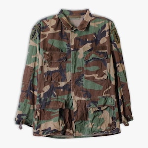 Könnte beinhalten: Tarnmusterjacke mit mehreren Taschen und einem Kragen. Die Jacke weist eine Mischung aus Grün-, Braun- und Schwarztönen auf. Auf den oberen Ärmeln sind Aufnäher zu sehen. Es handelt sich um ein Kleidungsstück im Militärstil.