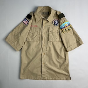 May include: Tan short-sleeved Boy Scouts of America shirt with two chest pockets and button closures. Features patches including the American flag, a Boy Scouts emblem, and a "Atlanta 1996" patch.