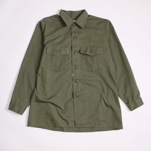 Puede incluir: Una camisa de manga larga en color verde oliva con cuello estándar, tapeta de botones frontal completa y dos bolsillos en el pecho con solapas abotonadas. La camisa tiene mangas largas con puños abotonados y un diseño de dobladillo recto.