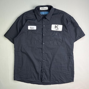 May include: Black short-sleeved work shirt with two chest pockets and a button-down front. The left pocket has a patch that says "Ruber", and the right pocket has a patch with a white "K" on a black background. A tag is visible at the collar.