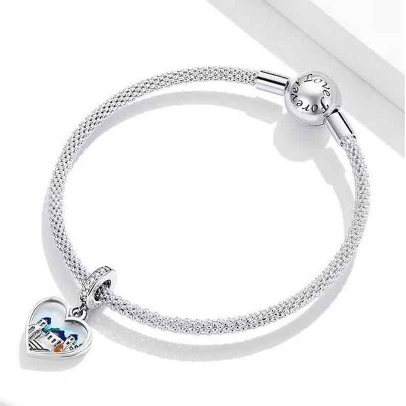 Puede incluir: Un brazalete de plata con un dije en forma de coraz&oacute;n que presenta una ciudad colorida. El brazalete tiene un cierre que dice "Love Forever".