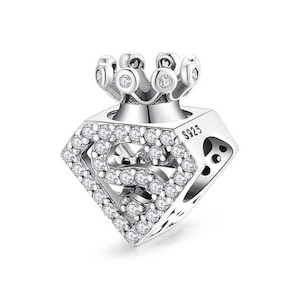 Könnte beinhalten: Silberner Anhänger mit einem Superman-Logo und einer Krone oben. Der Anhänger ist mit klaren Zirkonia-Steinen verziert. Der Anhänger ist seitlich mit "S925" gekennzeichnet.