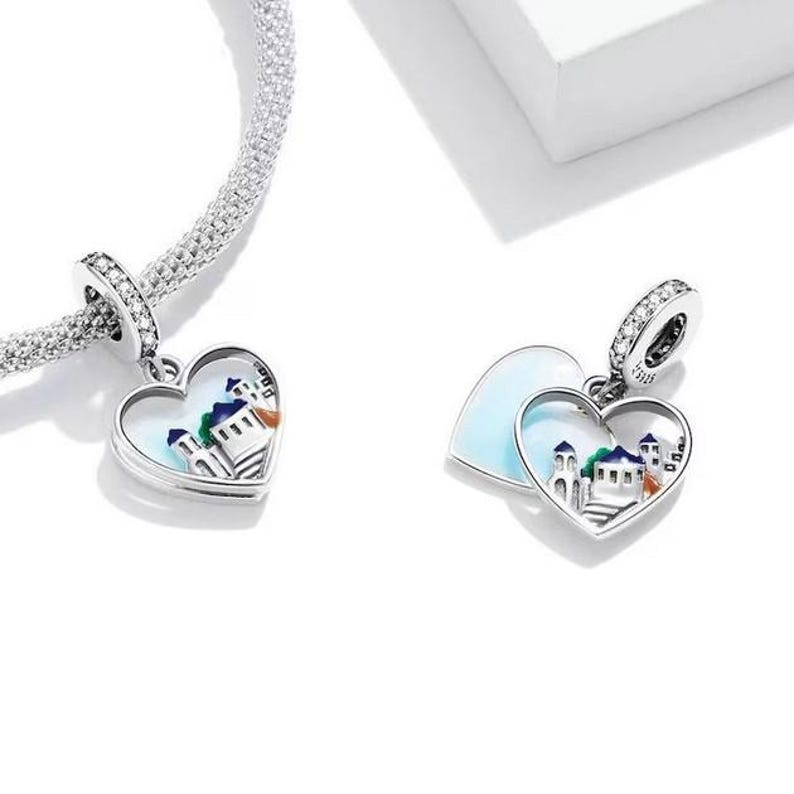 Puede incluir: Un charm de plata con dos corazones entrelazados. Los corazones est&aacute;n abiertos y revelan una escena pintada de un pueblo peque&ntilde;o con edificios y un cielo azul. El charm est&aacute; decorado con peque&ntilde;os cristales transparentes.