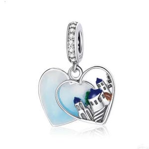 Può includere: Ciondolo in argento con due cuori intrecciati. Un cuore è in smalto blu e bianco pieno, l'altro è aperto e presenta un'illustrazione dettagliata di un piccolo paese con edifici bianchi e tetti blu.