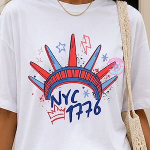 Pode incluir: Uma t-shirt branca de manga curta com um gráfico da coroa da Estátua da Liberdade em vermelho e azul. Abaixo da coroa, o texto diz NYC 1776 em azul, acompanhado por um pequeno ícone de coroa vermelha, estrelas azuis e raios vermelhos.
