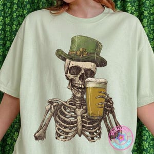 Op de afbeelding: Lichtgroen t-shirt met een afbeelding van een skelet met een groene hoed met een klaver en een glas bier. Het skelet lacht. De tekst "OH HAPPY PIXELS PNG" staat rechtsonder.