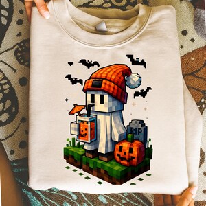 May include: A light beige sweatshirt featuring a pixelated Halloween-themed design. The graphic includes a ghost character wearing a red and orange hat, holding a drink with an orange slice, and surrounded by bats, a pumpkin, and a tombstone.