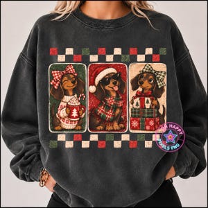 May include: A charcoal grey crewneck sweatshirt featuring a graphic print of three dachshunds. The dogs are depicted in three separate rectangular panels, wearing holiday accessories like bows, a Santa hat, and scarves, with mugs and gifts.