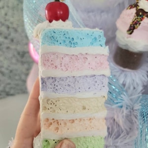 May include: A colorful layered cake ornament with a red cherry on top. The cake is decorated with a white frosting and has layers of blue, pink, purple, yellow, peach, and green. The ornament is hanging from a gold string.