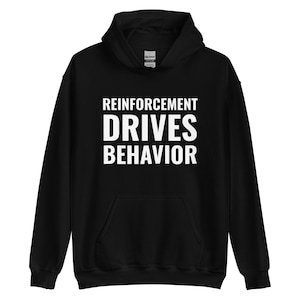 Puede incluir: Sudadera con capucha negra con el texto blanco "REINFORCEMENT DRIVES BEHAVIOR". La sudadera tiene un bolsillo delantero y una capucha con cordón. La etiqueta de la marca Gildan es visible en el cuello.