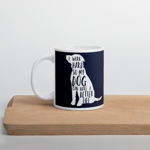 May include: White ceramic coffee mug with a blue background and a white silhouette of a dog. The text on the mug reads "I work hard so my dog can have a better life."