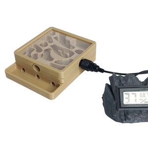 May include: A beige plastic ant farm with a clear plastic top and a black wire connected to a black and gray digital hygrometer that reads 68.7%.