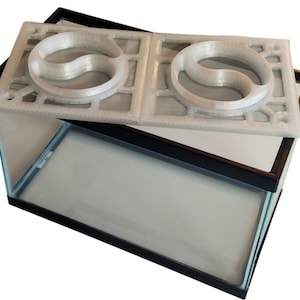 May include: A white plastic reptile habitat lid with two yin-yang shaped food dishes. The lid is sitting on top of a black framed glass tank.