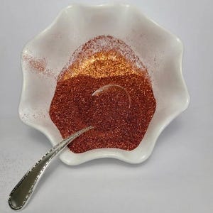 Op de afbeelding: Een witte keramische kom met een gegolfde rand gevuld met koperkleurige glitter. Een zilveren lepel rust in de kom, gedeeltelijk ondergedompeld in de glitter. De glitter gaat over van goud naar een diepe kopertint.