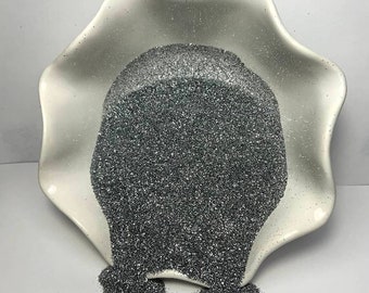 Puede incluir: Un cuenco de cerámica blanco con borde festoneado rebosante de purpurina gris oscuro. Parte de la purpurina se desborda por el lateral, creando un pequeño montón. La purpurina tiene una cualidad reflectante y brillante, lo que sugiere un material artesanal o decorativo.