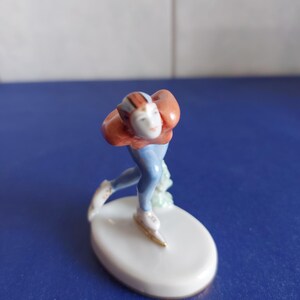 May include: A porcelain figurine of a speed skater in a forward-leaning posture. The skater wears a rust-orange top with dark stripes, light blue pants, and white skates. The figure is mounted on a smooth, oval white base with a small green accent.