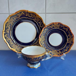 May include: A porcelain tea set consisting of a large plate, a smaller saucer, and a teacup. Each piece features a deep cobalt blue border with intricate gold filigree patterns and scalloped edges, surrounding a plain white center.