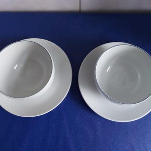 May include: Two white ceramic tea cups with matching saucers, each featuring a thin, dark blue rim around the top edge of the cup. The cups have a simple, rounded handle on the side and a smooth, plain white interior and exterior surface.