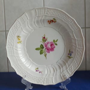 May include: White ceramic serving plate with a floral rose design. The plate has a scalloped edge with a textured pattern. A pink rose is in the center, with smaller floral accents around the rim. The plate is displayed on a clear stand.