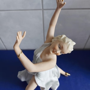 Może przedstawiać: Porcelanowa figurka baletnicy w pełnej gracji pozie. Figurka ubrana jest w białą sukienkę ze złotymi akcentami na nadgarstkach i stopach. Baletnica ma blond włosy i patrzy w dół z łagodnym wyrazem twarzy.