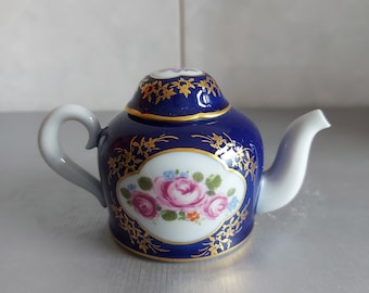 Puede incluir: Una tetera pequeña azul y dorada con un diseño floral. La tetera tiene una tapa y un pico. El diseño floral presenta rosas rosas y hojas verdes.