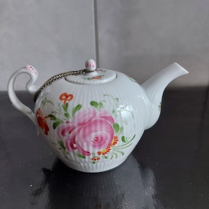 Può includere: Una teiera in porcellana bianca con un motivo floreale. La teiera presenta una rosa rosa, foglie verdi e fiori arancioni. La teiera ha una catena d'argento attaccata al coperchio.
