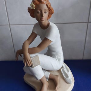 Può includere: Una statuetta in porcellana raffigurante una persona con capelli rossicci e un piccolo fiore giallo, che indossa una maglietta bianca e pantaloni bianchi. La figura è seduta, tiene un piccolo libro e ha un paio di scarpe bianche sulla base.