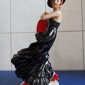 May include: A porcelain figurine of a woman in a black dress and red shoes, holding a red fan. She is wearing a black hat and has red hair.