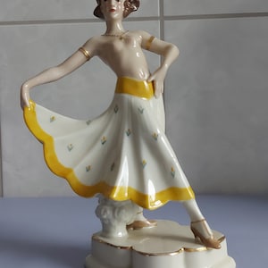 May include: A porcelain figurine of a woman in a white and yellow dress with a floral pattern. She is standing on a white pedestal with a gold trim.