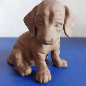 May include: A brown, matte-finished ceramic figurine of a seated puppy with long, drooping ears and a smooth, sculpted surface. The puppy is depicted in a sitting position with its head tilted slightly downward, showcasing detailed facial features.