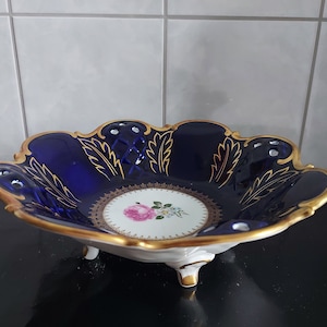 以下が含まれることがあります： 花柄と3本の足が付いた青と金の磁器のボウル。ボウルは金色の縁取りと中央の花柄で飾られています。