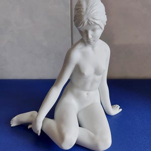 May include: A white porcelain figurine of a seated woman. The figure is kneeling, with arms extended and head bowed. The sculpture has smooth, detailed features and is set against a blue surface.