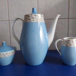 Könnte beinhalten: Vintage Teeservice aus Keramik in Hellblau, Weiß und Gold. Das Set besteht aus einer Teekanne, einem Sahnekännchen und einer Zuckerdose mit Deckel. Die Teekanne hat eine hohe, schlanke Form mit gebogenem Ausguss und Griff.