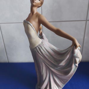 Peut inclure: Figurine en porcelaine d'une femme dans une robe fluide. La figurine est dans une pose gracieuse, un bras levé et l'autre tenant le bord de la robe. La robe est rose et blanche, et la figurine a une expression sereine.